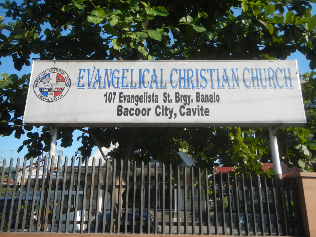 Eine evangelische Kirche in Bacoor City, Cavite, mit einem Schild, einem Metallzaun, einer Wand, Bäumen, Fahrzeugen, Gebäuden, Pfosten und einem bewölkten Himmel.