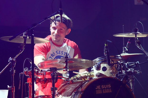 Ein Mann in einem roten und weißen T-Shirt spielt Schlagzeug auf der Bühne bei einem Konzert, lächelt und ist von Mikrofonen umgeben.