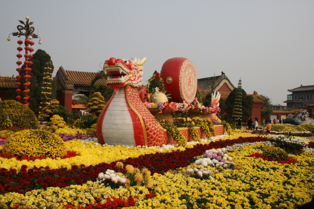 Ein farbenfroher chinesischer Neujahrsumzug zeigt eine Drachenstatue umgeben von bunten Blumen in Gelb, Rot, Pink und Weiß, mit Gebäuden, Bäumen und einem klaren blauen Himmel im Hintergrund, und Menschen, die um die festliche Szenerie herumgehen.
