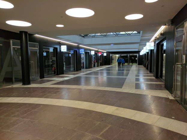 Ein langer Flur mit Menschen, die gehen, beleuchtet von Deckenlampen, umgeben von Säulen und Glastüren und mit Schildern mit Text, die den Eindruck eines belebten Rathauses vermitteln.