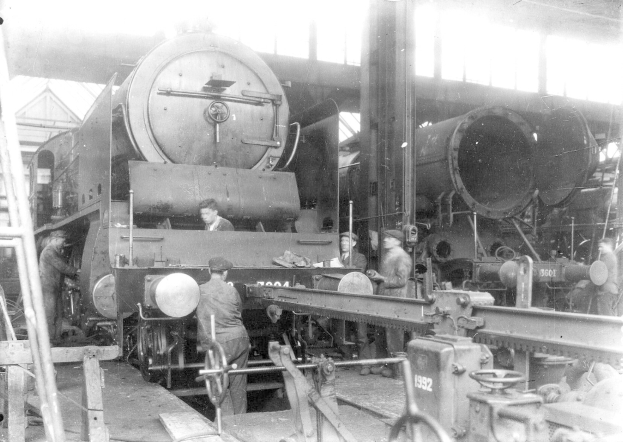 Schwarzes und weißes Foto von Arbeitern, die einen Zug in einer Fabrik reparieren, wobei einige Werkzeuge und eine Leiter auf der linken Seite zu sehen sind; im Hintergrund befindet sich eine Hütte.