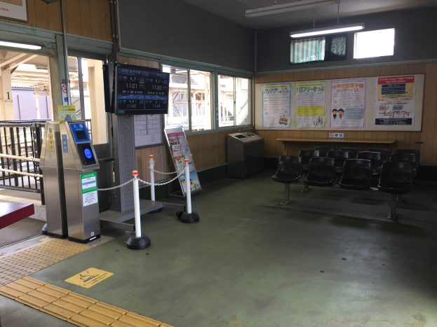 Ein belebter Bahnhofseingang mit Sitzgelegenheiten, einem Service-Schalter, Ticketautomaten, Deckenleuchten, Geländern, Informationsanzeigen und Wandplakaten.