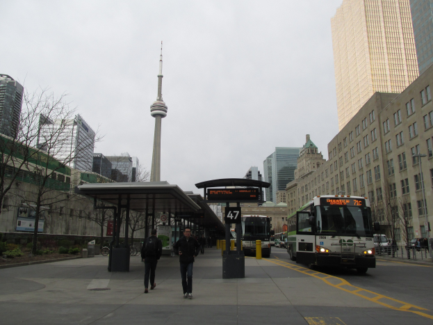 Eine belebte Stadtstraße mit einem Bus an einer Bushaltestelle vor einem hohen Gebäude, Fahrzeuge auf der Straße, Fußgänger auf dem Gehweg, ein überdachter Schuppen, Bäume, Gebäude mit Fenstern, ein Turm und der Himmel.