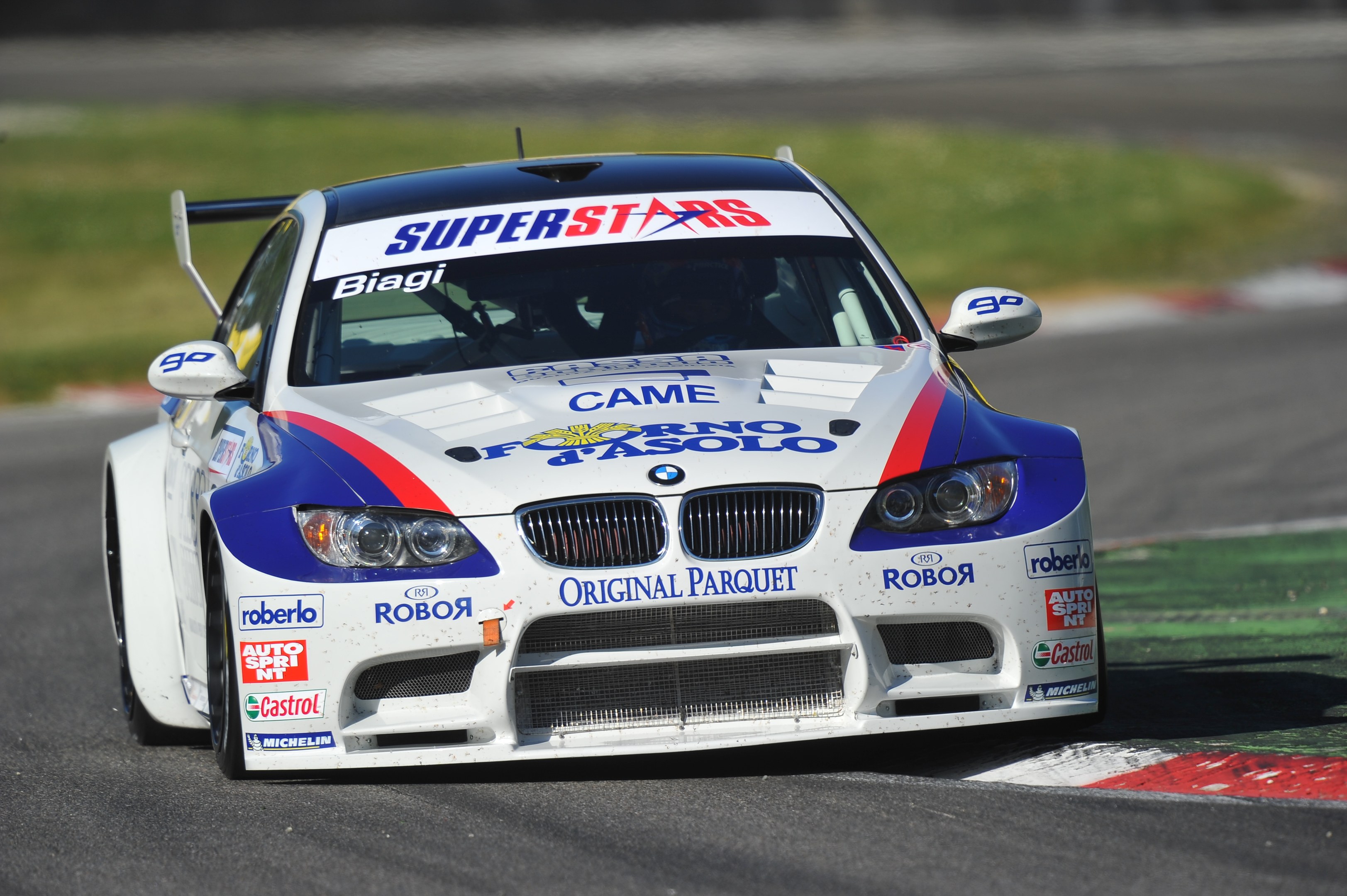 Ein BMW M3 GT3 Rennauto fährt auf einer unscharfen Rennstrecke mit Gras im Hintergrund.