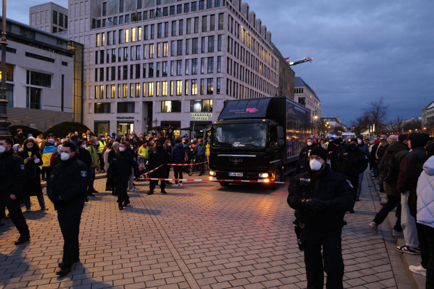 Eine Gruppe von Menschen steht in der Nähe eines Lastwagens auf einer Straße mit Gebäuden, Bäumen und Laternen im Hintergrund; einige tragen Mützen und Masken, und ein Band ist an einem Pfahl im Vordergrund befestigt.