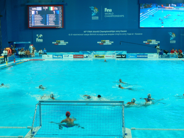 Eine Gruppe von Menschen spielt Wasserball in einem großen Pool mit einem Netz in der Mitte, Zuschauern auf der linken Seite und einer Wand mit einem Bildschirm im Hintergrund bei den FINA-Weltmeisterschaften 2016 in Kazan.