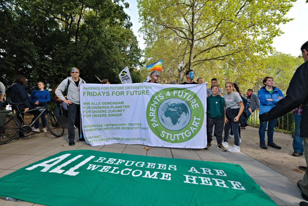 Gruppe von Menschen mit einem Banner "All Refugees Are Welcome Here" und einer Fahne, mit Fahrrädern, einem Zaun, einem Straßenschild, einer Tafel, Bäumen und einem bewölkten Himmel im Hintergrund.
