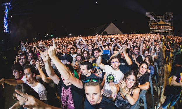 Menschenmenge auf einem Musikfestival mit erhobenen Händen, einige sitzen und andere stehen, umgeben von einem Metallzaun mit Gebäuden und Lichtern im Hintergrund.