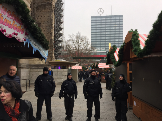 Polizeibeamte in schwarzen Uniformen und Mützen stehen vor einem geschmückten Weihnachtsmarkt in Berlin, mit festlichen Dekorationen, Gebäuden, Bäumen und einem klaren blauen Himmel im Hintergrund.
