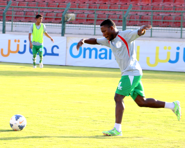 Ein Mann kickt einen Fußball auf einem Rasenfeld in einem Stadion mit zwei Personen in der Nähe und Bannern, Geländern und Stühlen im Hintergrund.