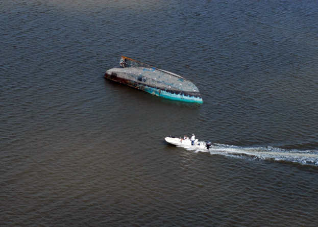 Ein kleines, teilweise untergetauchtes Wrackboot fährt einen Fluss hinunter neben einem größeren Boot mit ein paar Menschen an Bord.