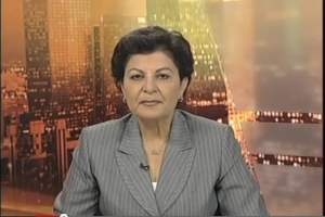 Eine Frau in einem Business-Outfit sitzt vor einem Fernseher, im Hintergrund sind ein paar Gebäude zu sehen, die anscheinend eine Nachrichtensprecherin zeigt, wie der Text am unteren Bildschirmrand anzeigt.