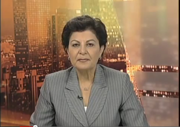 Eine Frau in einem Business-Outfit sitzt vor einem Fernseher, im Hintergrund sind ein paar Gebäude zu sehen, die anscheinend eine Nachrichtensprecherin zeigt, wie der Text am unteren Bildschirmrand anzeigt.