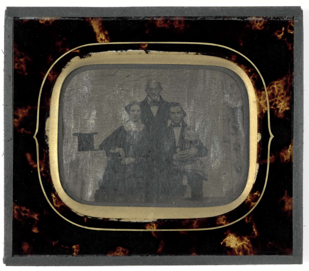 Schwarze-weiße Fotografie eines Mannes in Anzug und Krawatte und einer Frau in unbekannter Kleidung, die vor einem alten Fernseher stehen und ernst schauen.