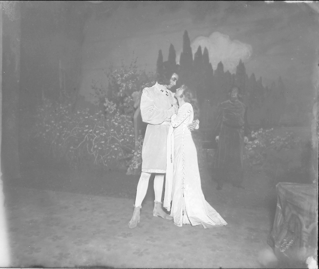 Ein Schwarz-Weiß-Foto von einem Mann und einer Frau, die sich auf einer Bühne umarmen und küssen, mit einem Publikum, Pflanzen und Bäumen im Hintergrund.