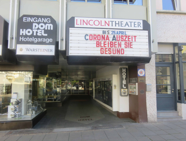 Außenansicht des Lincoln Theaters in Berlin, Deutschland, mit Glasfenstern, Türen und einer Texttafel, wobei im Inneren durch die Fenster eine belebte Stadtlandschaft zu sehen ist.