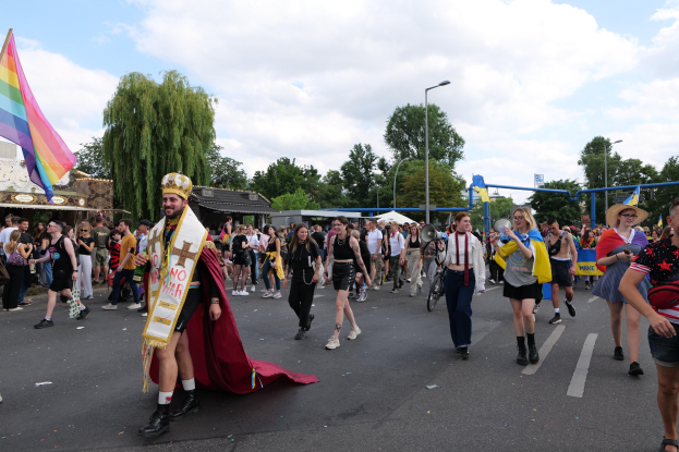 Eine Gruppe von Menschen bei der 2018 Gay Pride Parade mit einer Regenbogenfahne und Musikinstrumenten, einige tragen Mützen, vor dem Hintergrund von Laternenpfählen, Bäumen, Schuppen und einem bewölkten Himmel.