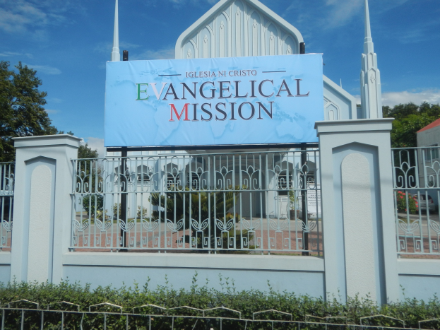 Kirche mit Metallzaun, Schild mit der Aufschrift "Evangelische Mission", umgeben von Bäumen, einem Haus mit Dach und einem bewölkten Himmel.