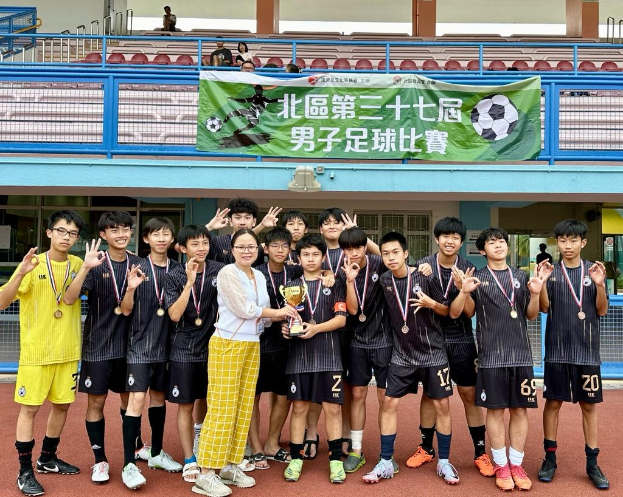 Gruppe junger Männer in Fußballtrikots auf einem Feld, die Medaillen tragen und einen Pokal halten, mit einem Banner für die "Yokohama U-16 Jungenfußballmannschaft" im Hintergrund und sitzenden Zuschauern.