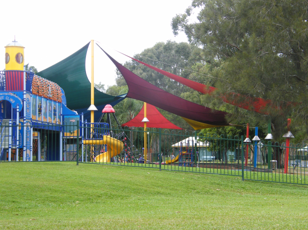 Ein Park mit einem Spielplatz, der von einem Metallzaun umgeben ist und Kinder auf Geräten wie einer Rutsche, Pfosten und einem Zelt spielt, umgeben von grünem Gras, Bäumen und einem klaren blauen Himmel.