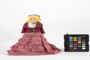 Eine Barbie-Puppe in einem pinken Kleid und einer maronenfarbenen Jacke steht neben einem schwarzen Farbpaletten auf einer weißen Oberfläche.