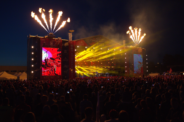 Eine große Menschenmenge versammelt sich auf einem Musikfestival mit einer Bühne, auf der Musiker unter hellen Scheinwerfern spielen und von Bannern umgeben sind, links davon Zelte und im Hintergrund ein dunkler Himmel mit Feuerwerk.