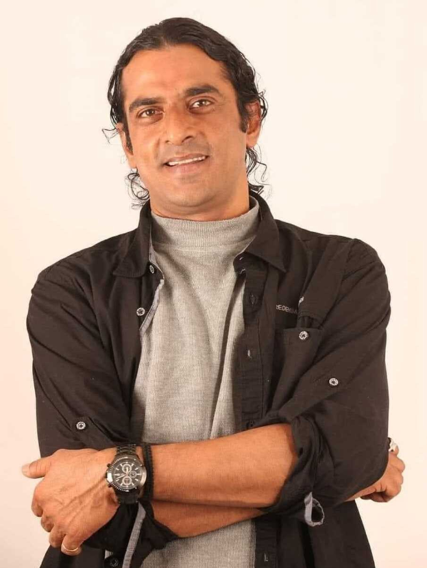 Ein Mann in einem schwarzen und aschfarbenen Kleid mit einer Uhr steht mit verschr├Ąckten Armen tief in Gedanken vor einem wei├čen Hintergrund.