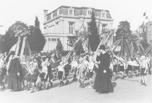 Schwarz-weißes Bild einer Gruppe von Menschen, die eine Straße entlanggehen und Fahnen vor einem Gebäude halten, mit Bäumen und anderen Gebäuden im Hintergrund während einer Mai-Feier.