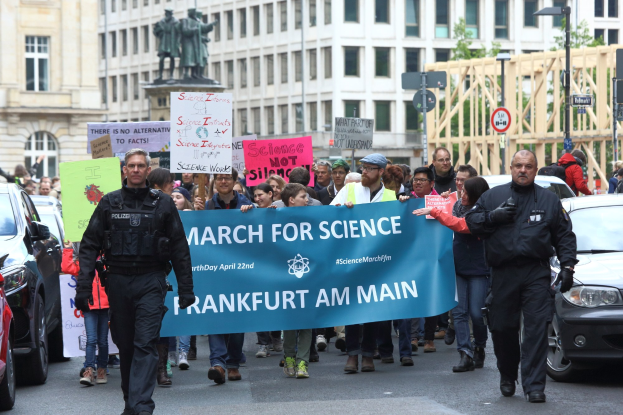 Eine Gruppe von Menschen marschiert mit einem "March for Science Frankfurt am Main"-Schild die Straße entlang, mit Autos, Gebäuden, Statuen und Bäumen im Hintergrund.
