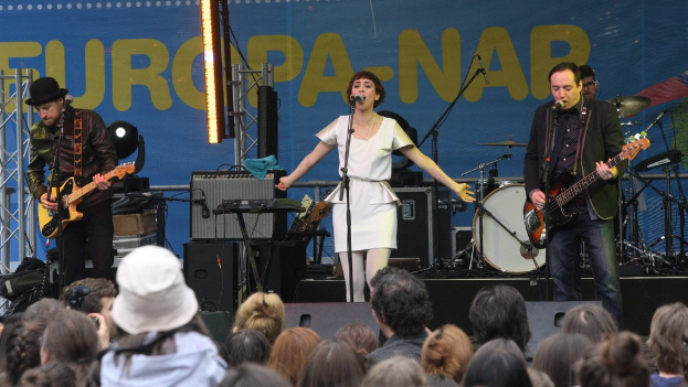 Vier Musiker auf einer Bühne: Eine Frau in einem weißen Kleid singt in der Mitte, flankiert von zwei Männern mit Gitarren, während das Publikum unten zusieht, mit Lautsprechern und einer Wand im Hintergrund.