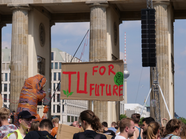 Eine Gruppe von Menschen steht vor dem Brandenburger Tor in Berlin, Deutschland, wobei einige Taschen tragen und einer im Kostüm mit einer Tafel steht; Gebäude, Bäume und ein Turm sind im Hintergrund unter einem bewölkten Himmel zu sehen.