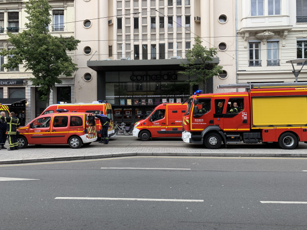 Eine Gruppe von Feuerwehrautos auf einer Pariser Straße mit Menschen in der Nähe, vor Gebäuden, Bäumen und einem Fahrrad, wahrscheinlich nach einem Unfall.
