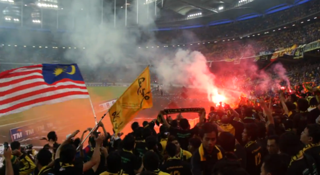 Eine große Menschenmenge in einem Stadion mit Bengalos, Fahnen und einer Kamera, mit Rauch und Schildern, die 'Malaysia vs Malaysia Live Streaming' lesen.