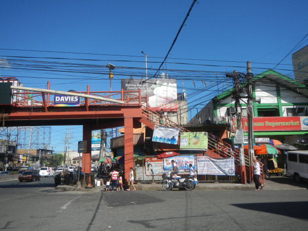 Eine belebte Stadtstraße mit einer Brücke darüber, Fahrzeuge, Fußgänger, Strommasten, Gebäude, Banner, Straßenlaternen, Bäume und einen klaren blauen Himmel.