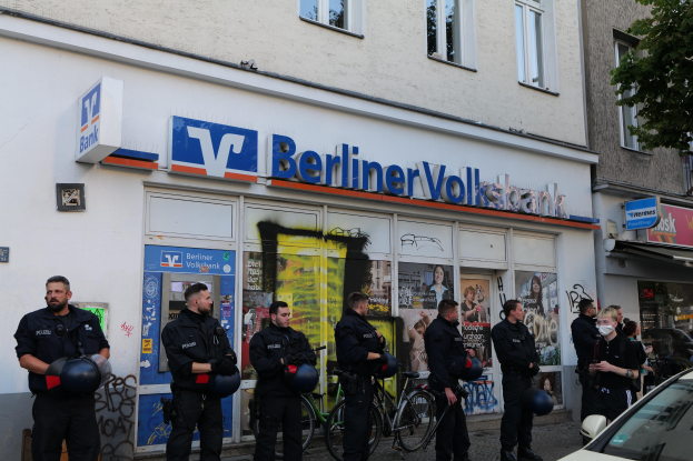 Gruppe von Polizeibeamten vor einer Berliner Volksbank, einige halten Helme, rechts ein Auto und links ein Baum, Gebäude und Schilder im Hintergrund.