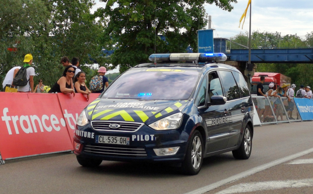 Polizeiauto fährt auf einer Straße neben einer Menschenmenge, mit Bannern, Geländern, Bäumen, einer Brücke, einer Flagge und einem bewölkten Himmel im Hintergrund.