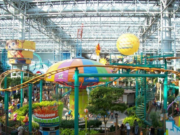 Ein großes Indoor-Wasserpark mit einer Achterbahn, umgeben von Menschen, Bäumen, Pflanzen und einer farbenfrohen Beleuchtung von oben.