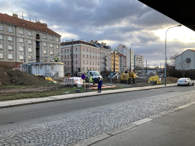 Eine Straße in der Stadt mit parkenden Autos, Gebäuden, Laternen, Bäumen und einem bewölktem Himmel, mit einer Sperre mit Fahrzeugen und Füßgängern im Vordergrund.