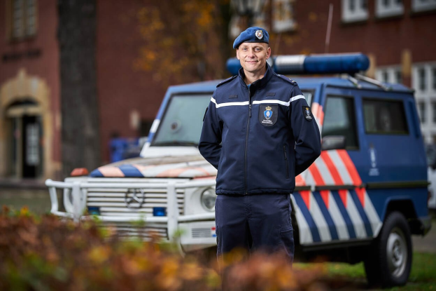 Polizist in Uniform vor einem Polizeiwagen mit leicht unscharfem Hintergrund und Pflanzen unten.