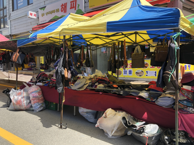 Ein Straßenmarkt mit Zelten, die verschiedene Gegenstände wie Taschen und Hüte präsentieren, vor Häusern mit Fenstern und Schildern im Hintergrund.