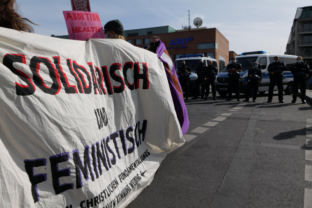 Eine Gruppe von Menschen marschiert auf einer Straße, die eine 'Solidarität und Feminismus'-Tafel hält, mit geparkten Fahrzeugen und Gebäuden im Hintergrund unter einem klaren blauen Himmel.