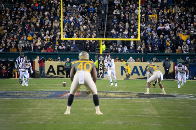 Ein Fußballspiel wird in einem Stadion mit Spielern in Sportbekleidung und Helmen im Vordergrund und einer Zuschauermenge im Hintergrund sowie sichtbaren Pfosten und Tafeln mit Text gespielt.