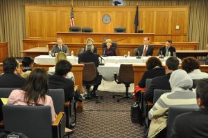 Ein Versammlungsraum mit einer Gruppe sitzender Menschen, zwei ausländische Flaggen, ein oranger Schreibtisch mit Wasserflaschen und einem Mikrofon.