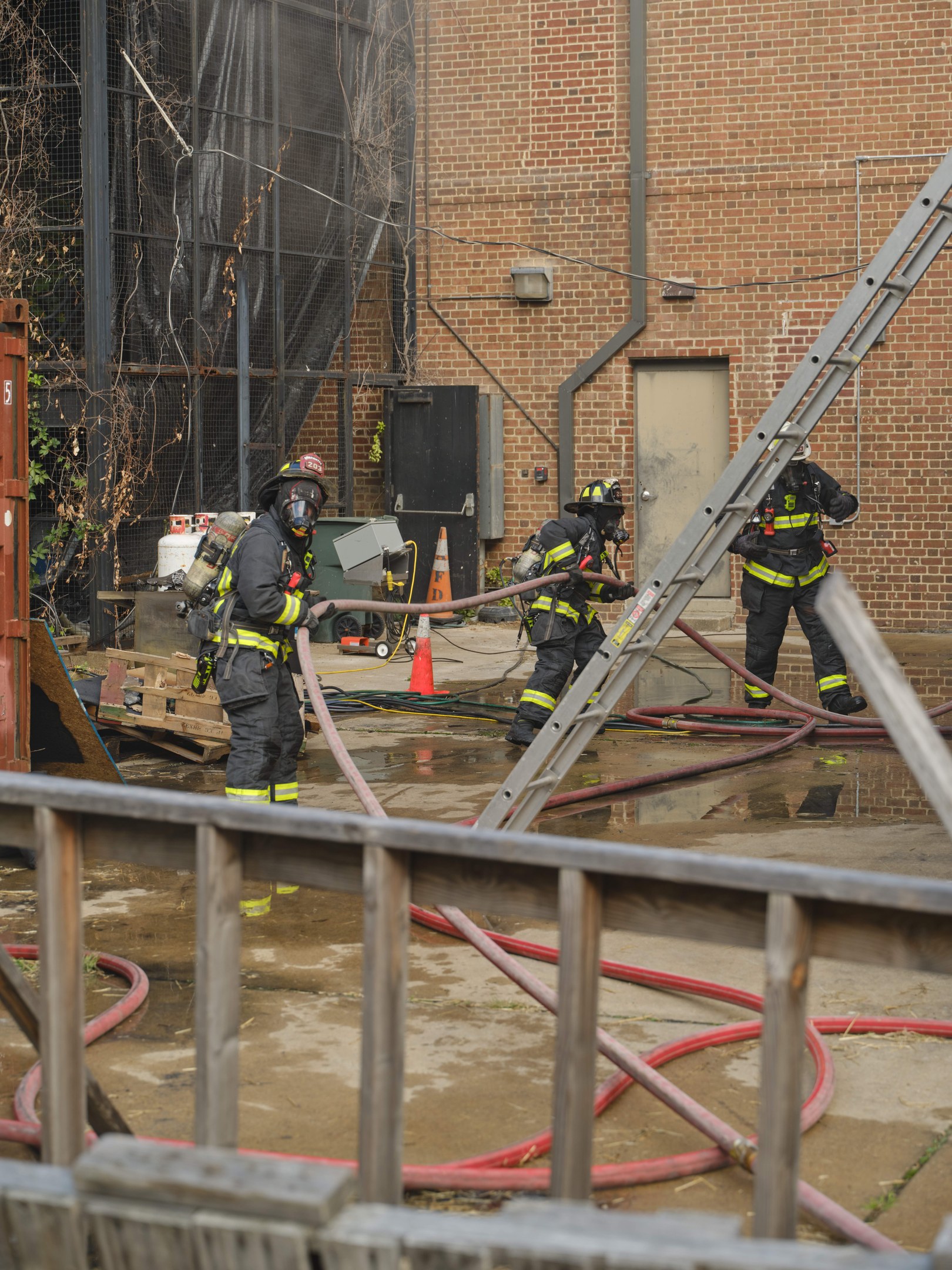 Feuerwehrleute in Helmen arbeiten daran, ein Gebäude Feuer zu löschen, während sie Schläuche halten, mit einem Metallzaun, verstreuten Rohren, einem Container, einem Verkehrskegel und Hintergrundelementen einschließlich eines Baumes und des Himmels.