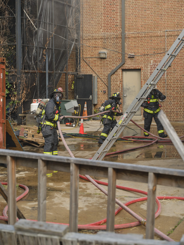 Feuerwehrleute in Helmen arbeiten daran, ein Gebäude Feuer zu löschen, während sie Schläuche halten, mit einem Metallzaun, verstreuten Rohren, einem Container, einem Verkehrskegel und Hintergrundelementen einschließlich eines Baumes und des Himmels.