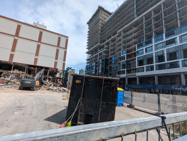 Großes Gebäude mit umfangreichem Schutt davor, umgeben von einem Metallzaun, neben einem Container, Pflanzen, Bäumen, Pfählen, einem Schild, Fahrzeugen, Steinen und einem bewölkten Himmel, was auf eine Baustelle für ein neues Wohngebäude hinweist.