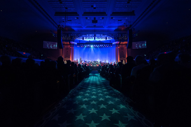 Ein großer Saal mit sitzenden Zuschauern, eine Bühne mit Musikern und eine Decke mit Lampen und Fernsehgeräten, was auf ein Konzerthaus während der Nationalkonvention 2019 hindeutet.