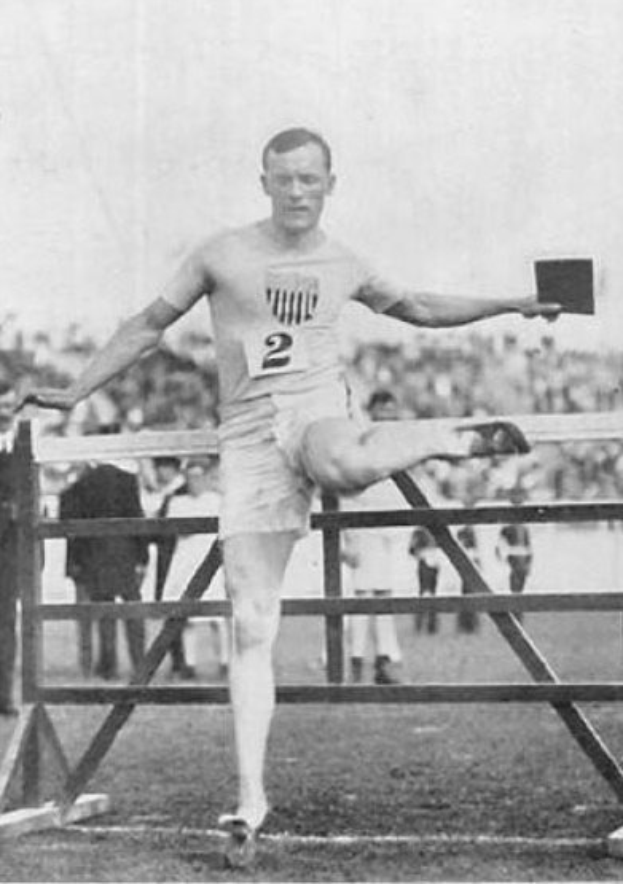 Schwarz-weißes Foto eines Mannes in Sportkleidung, der über ein Hindernis springt, mit einer Zahl auf Hemd und Shorts, vor Zuschauern unter freiem Himmel.