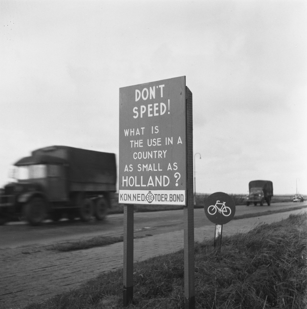 Schwarz-weißes Foto eines Lkw, der auf einer Straße mit einem "Fahr nicht zu schnell"-Schild im Vordergrund und anderen Fahrzeugen, Gras, Pfosten und einem klaren Himmel im Hintergrund fährt.