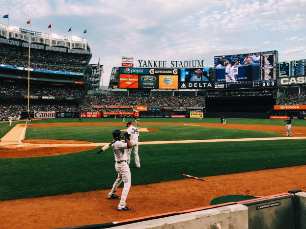 Baseballspiel im Yankee Stadium mit Spielern und Zuschauern, umgeben von Stadionmerkmalen wie Zäunen, Flaggen, Anzeigetafeln, einem Bildschirm und Deckenlampen unter einem bewölkten Himmel.
